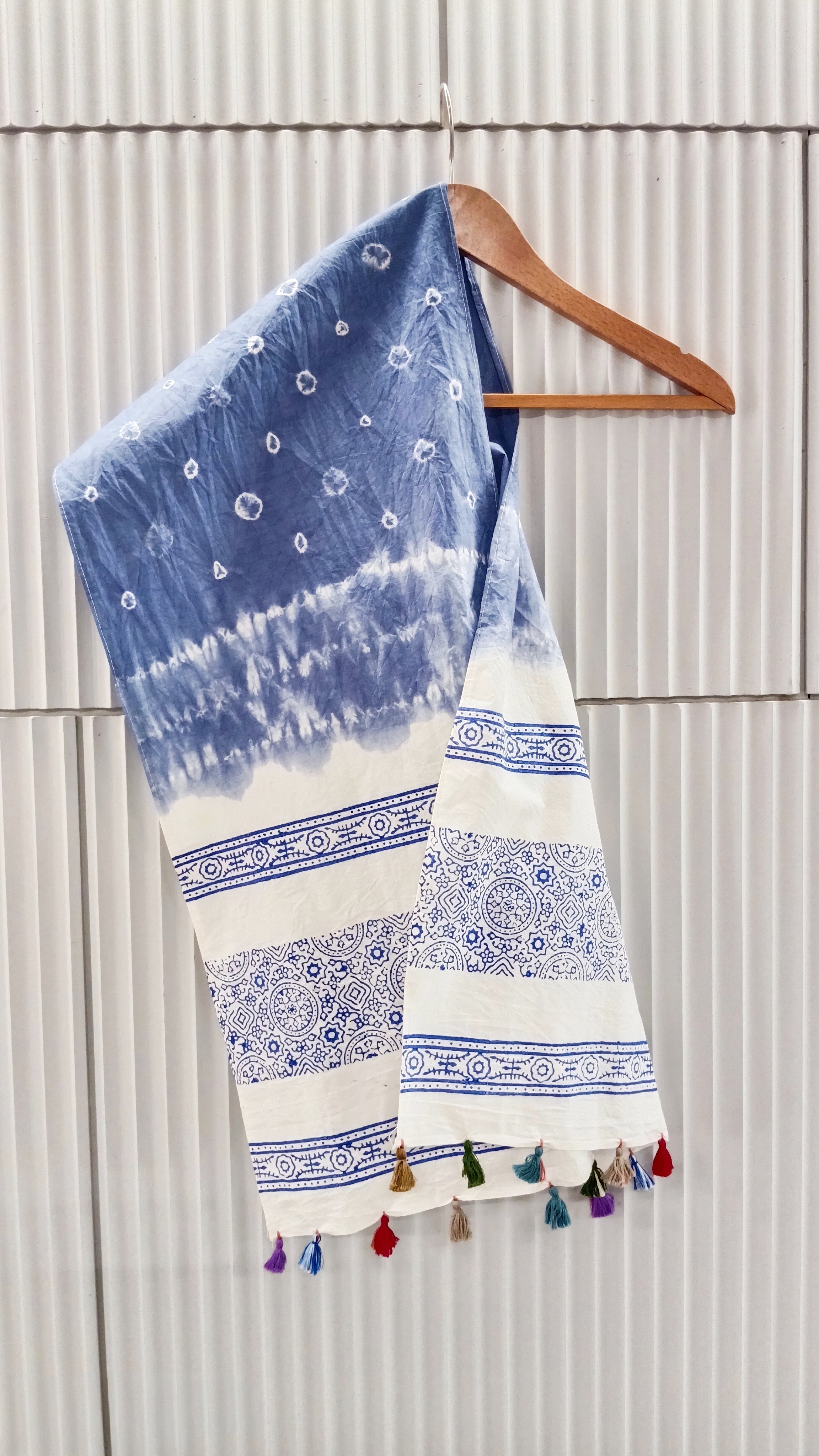 Blue and white patterned stole with tassels on a wooden hanger against a light gray wall.